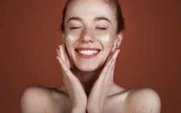 Smiling caucasian ginger lady with freckles applying anti aging cream on face and smile posing with naked shoulders on red studio wall