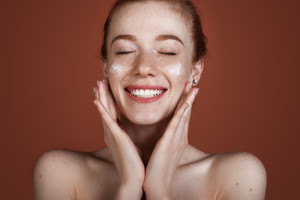 Smiling caucasian ginger lady with freckles applying anti aging cream on face and smile posing with naked shoulders on red studio wall